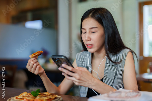 Billede på lærred Asian woman sitting working in cafe, With relaxation, take break from work listen music by using headphones connected phone, picked up pizza from tray ate it deliciously, happy face when eating