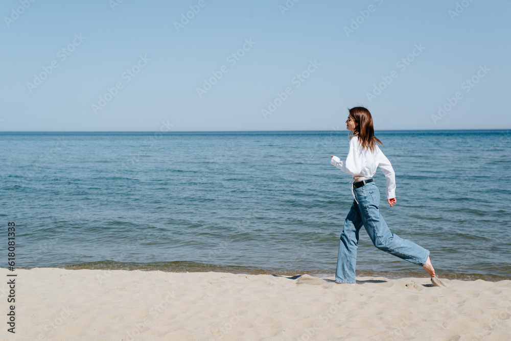 Lifestyle, freedom, carelessness. Barefoot young woman in sunglasses and casual clothes running on seashore on sunny day, copy space