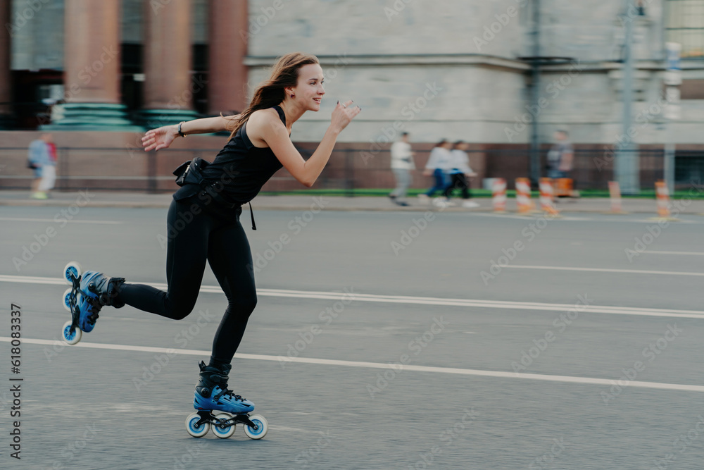 Holidays and active lifestyle concept. Slim healthy European woman rollerskates on high speed photogaphed in motion dressed in black clothing poses against blurred street background on road.