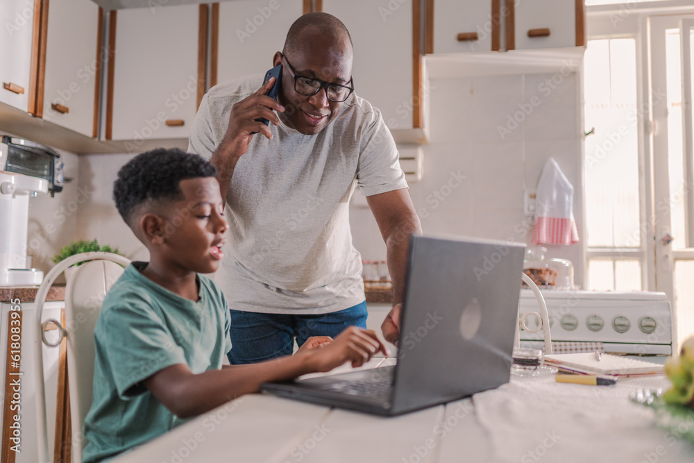 pai Latino ajudando seu filho a usar o computador portatil para fazer a tarefa da escola enquanto fala ao celular sobre trabalho na cozinha de sua casa no Brasil pai Latino ajudando seu filho a usar o computador portatil para fazer a tarefa da escola enquanto fala ao celular sobre trabalho na cozinha de sua casa no Brasil