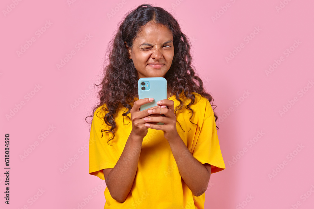Young funny Indian woman teenager with phone in hands closes one eye coming up with response to friend messages or comment in social network stands on isolated pink background. SMS, Email, chatting Young funny Indian woman teenager with phone in hands closes one eye coming up with response to friend messages or comment in social network stands on isolated pink background. SMS, Email, chatting