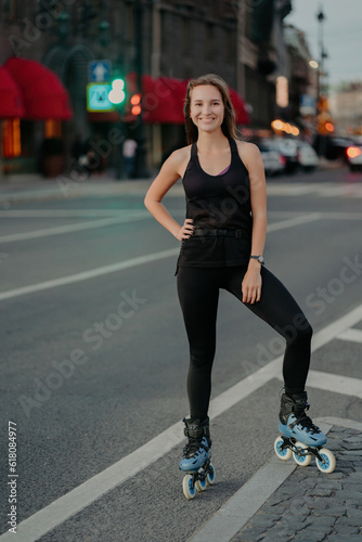 Pleased Eurpean woman keeps hand on waist smiles pleasantly leads active lifestyle rides rollerskates dressed in black clothing poses against blurred busy city background. Extreme sport concept