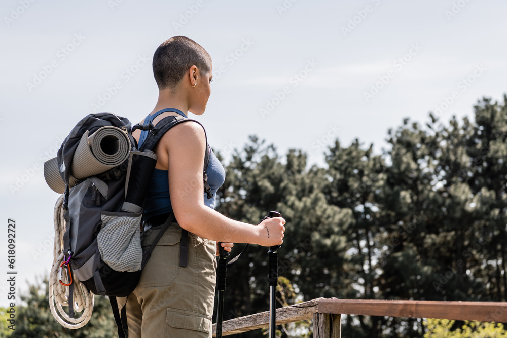 Young short haired woman tourist in casual clothes with backpack and travel equipment holding trekking poles while standing with nature at background, Translation of tattoo: love