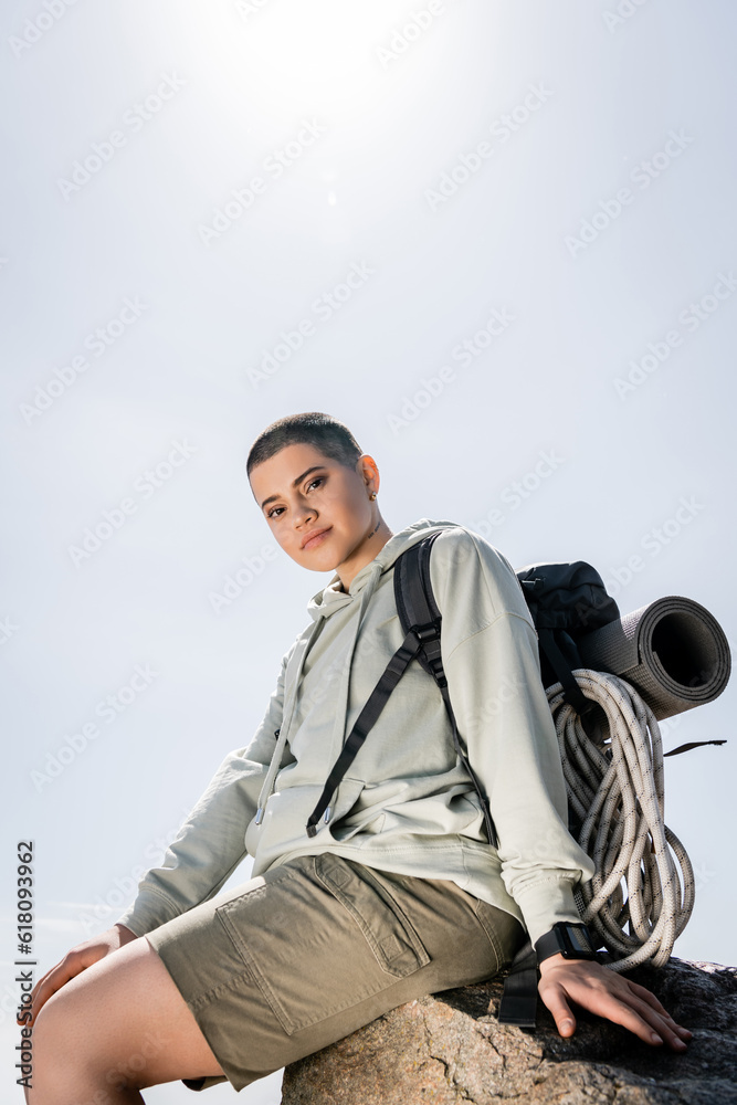 Low angle view of young short haired and tattooed woman hiker with fitness tracker and backpack looking at camera while sitting on stone with blue sky at background, vibrant travel experiences Low angle view of young short haired and tattooed woman hiker with fitness tracker and backpack looking at camera while sitting on stone with blue sky at background, vibrant travel experiences