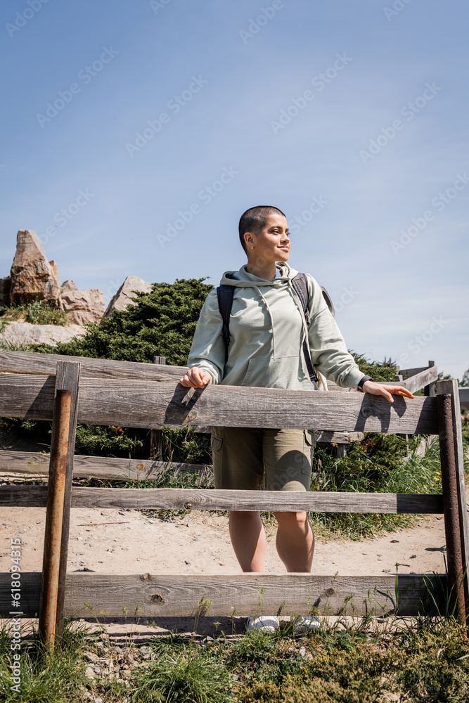 Fototapeta premium Smiling young short haired female traveler with backpack in casual clothes looking away while standing near wooden fence with nature at background, hiker trekking through landscape, summer