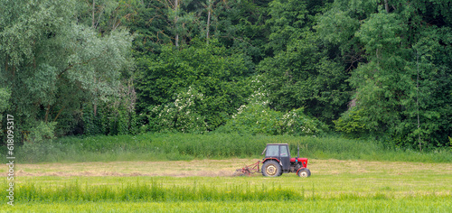 Zgrabianie siana za pomocą zgrabiarki przyłączonej do czerwonego traktora, na łące u stóp wzgórza porośniętego lasem liściastym . photo