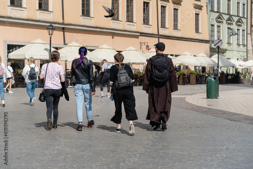 Authentic young priest in a black cassock walks with three teenage girls and backpacks along the street of the old European city of Krakow, a sunny summer day in city, multiculturalism and diversity