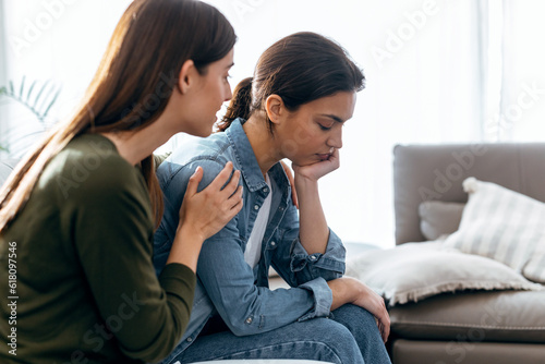 Bild auf Leinwand Pretty young woman supporting and comforting her sad friend while sitting on the sofa at home