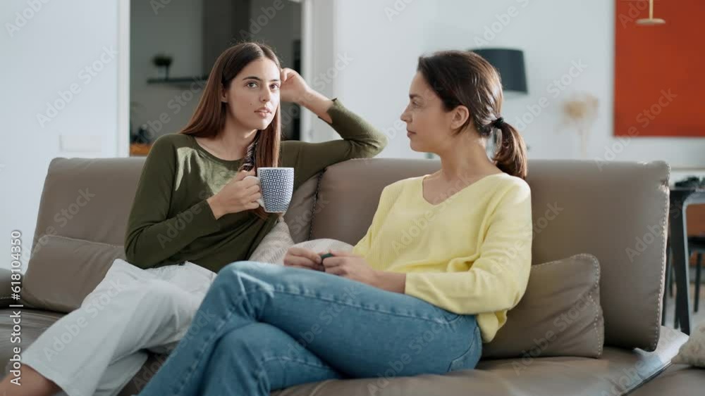 Video of two smiling young women talking while drinking coffee sitting on couch in the living room at home. Video of two smiling young women talking while drinking coffee sitting on couch in the living room at home.
