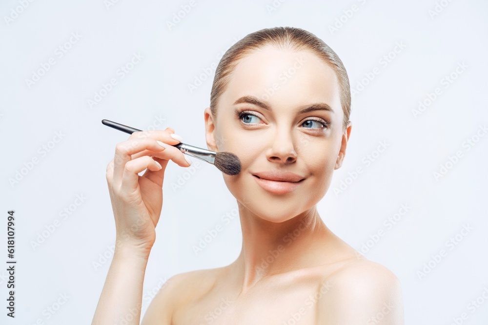 Facial treatment, cosmetology. Young woman, dark hair, applies powder with brush, sideways, naked body