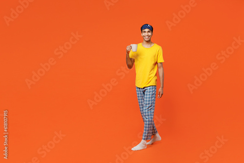 Full body smiling happy fun young man wears pyjamas jam sleep eye mask rest relax at home hold in hand cup of coffee isolated on plain orange background studio portrait. Good mood night nap concept.