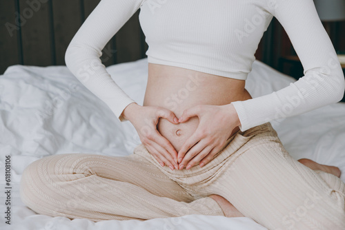 Cropped close up young pregnant woman wear white pyjamas hold hnds in heart shape belly rest relax spend time in bedroom lounge home in own room hotel. Maternity family pregnancy expectation concept. © ViDi Studio