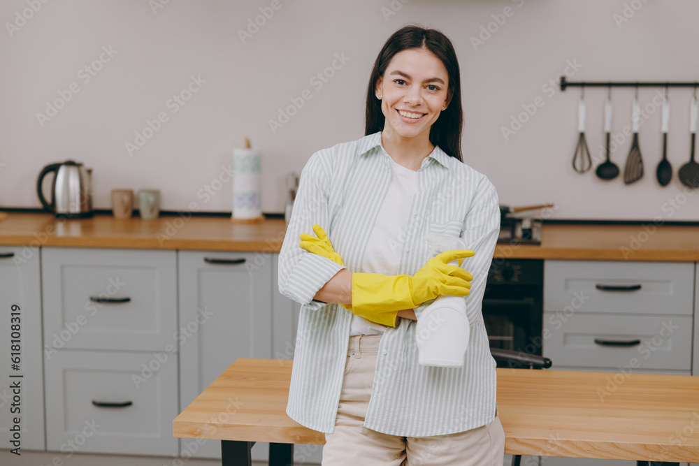 Young smiling happy housewife latin woman wearing casual clothes shirt hold detergent bottle while doing housework tidy up in light kitchen at home alone. Lifestyle cooking food housekeeping concept. Young smiling happy housewife latin woman wearing casual clothes shirt hold detergent bottle while doing housework tidy up in light kitchen at home alone. Lifestyle cooking food housekeeping concept.