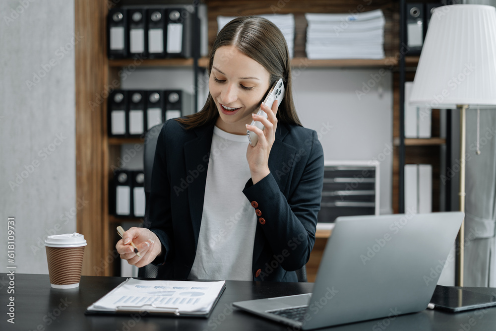 businesswoman talking mobile phone and working on laptop and papers on table in her office. businesswoman talking mobile phone and working on laptop and papers on table in her office.