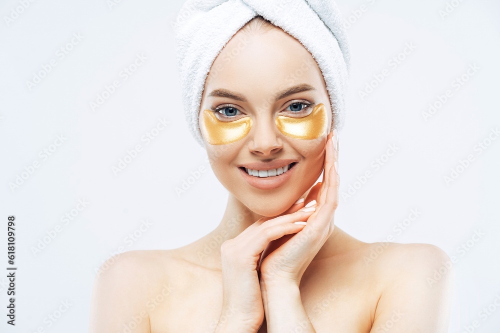 Pretty lady, towel-wrapped head, touches cheeks, applies golden hydrogel patches, shirtless indoors. Removes wrinkles, healthy skin Pretty lady, towel-wrapped head, touches cheeks, applies golden hydrogel patches, shirtless indoors. Removes wrinkles, healthy skin