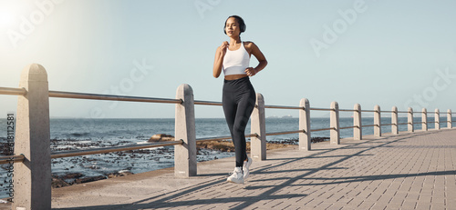 Fitness, woman run at beach and with headphones listening to music for health wellness. Training or exercise, marathon or lens flare and female person running along the promenade listen to radio