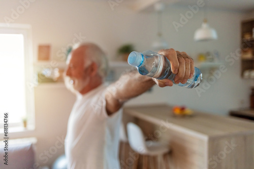 Smiling senior man exercising with a two bottle of water at home. Gray hair mature adult man exercising at home with hand weights in his living room.