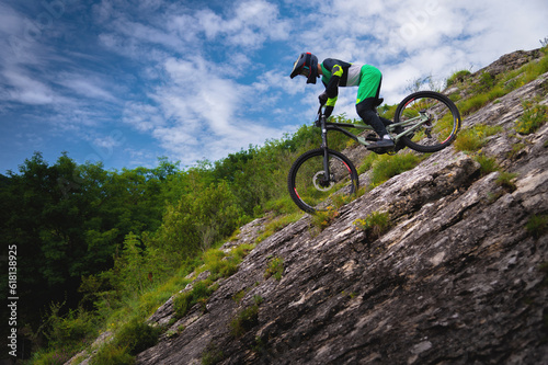 Downhill rider cyclist rides down a steep cliff surrounded by green trees against a blue sky. Copy space mtb concept