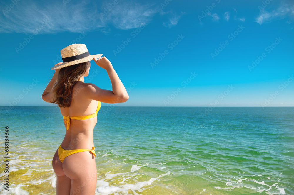 Attractive caucasian young woman in a yellow swimsuit stands with her back holding on to a straw hat and looks at the sea on a sunny day