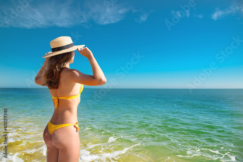 Attractive caucasian young woman in a yellow swimsuit stands with her back holding on to a straw hat and looks at the sea on a sunny day