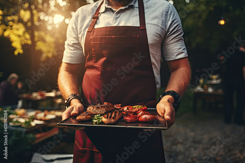 Obraz na płótnie A man in a red apron holds a tray with meat and vegetables on the background of a barbecue party