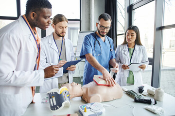 professional paramedic tamponing wound on simulator near young multiethnic students in white coats and medical equipment during first aid training seminar, effective life-saving skills concept