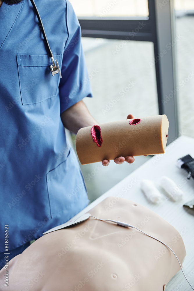 Obraz premium cropped view of professional paramedic in blue uniform holding wound care simulator near CPR manikin and bandages on blurred background in training room, critical skills development concept