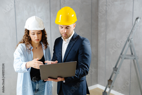 Attractive young woman contractor is talking to customer in helmet, and demonstrates sequence of repairs in living room on laptop. Meeting of landman of apartment with engineer or architect. photo