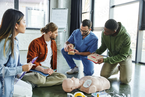 medical instructor and african american man holding wound care simulators near CPR manikin, defibrillator and multiethnic participants with notebook and clipboard, life-saving skills concept