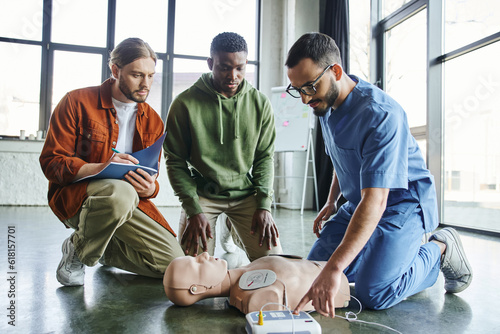 cardiac resuscitation, young man writing in notebook near african american participant and medical instructor operating defibrillator on CPR manikin during first aid training seminar photo