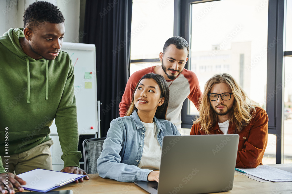 impressed multiethnic men looking at laptop near smiling asian woman, diverse group of young and creative business people working on startup planning in contemporary office