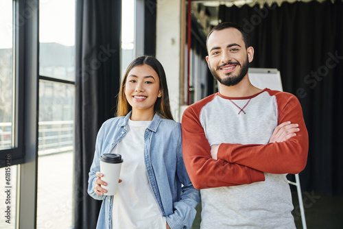 pleased asian woman with coffee to go in paper cup and bearded man with folded arms smiling at camera in modern office, young interracial entrepreneurs collaborating in startup