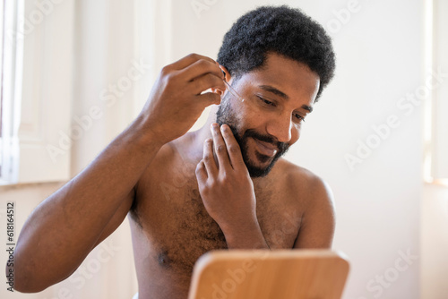Young bearded African American young man looking at himself in a small wooden mirror, and applying moisturising face oil for healthy nourished skin, skin care, hygiene concept