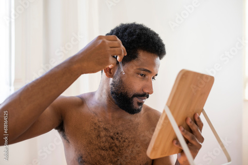 Young bearded African American young man looking at himself in a small wooden mirror, and applying moisturising face oil for healthy nourished skin, skin care, hygiene concept