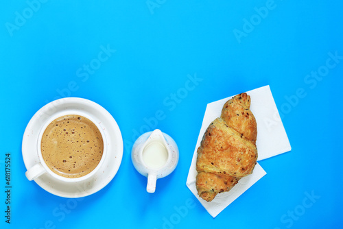 Fototapeta Naklejka Na Ścianę i Meble -  Cup with cappuccino, cereal croissant and milk jug, top view. Coffee and croissant on a blue background. The concept of healthy eating and sweet food. French breakfast. Flat lay. Background for text