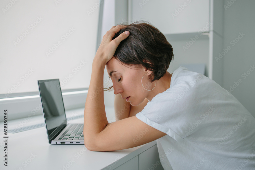 Tired, frustrated, exhausted, stressed woman sitting at desk working on laptop. Headache. Daylight work. Girl in White tshirt Tired, frustrated, exhausted, stressed woman sitting at desk working on laptop. Headache. Daylight work. Girl in White tshirt
