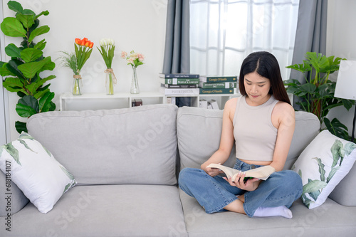 Young Asian woman reading a book at her home sitting on the fabric sofa In the living room having a good time at home
