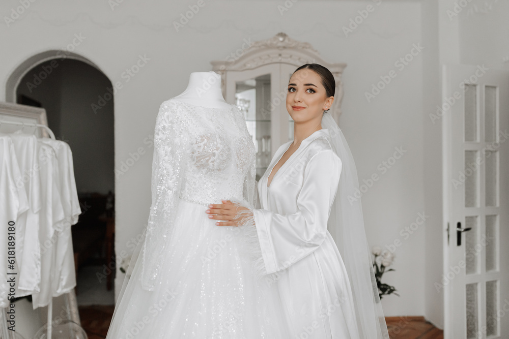 Stylish bride with wedding hairstyle and makeup in white robe standing near her wedding dress. Stylish bride with wedding hairstyle and makeup in white robe standing near her wedding dress.