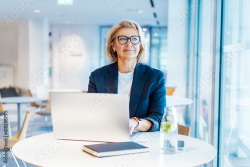 50's confident mature businesswoman dreamy looking at window, middle-aged experienced senior female professional working on laptop in open space office. Female entrepreneur working remotely
