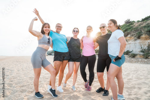 Cheerful sporty group of active women of various ages smiling at camera together standing on the beach after workout training. Female companions in sport resting after fitness. Health and wellbeing.