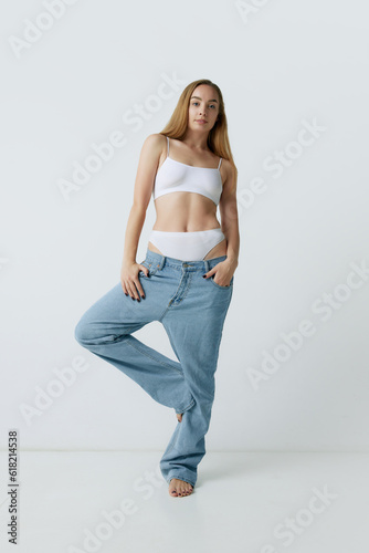 Full-length portrait of beautiful young blonde woman with slim body posing in jeans and top against grey studio background. Concept of beauty, body and skin care, health, fitness, wellness, ad