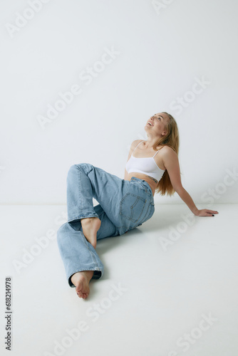 Beautiful, smiling, young blonde woman with slim body posing in white top and jeans against grey studio background. Concept of beauty, body and skin care, health, fitness, wellness, ad