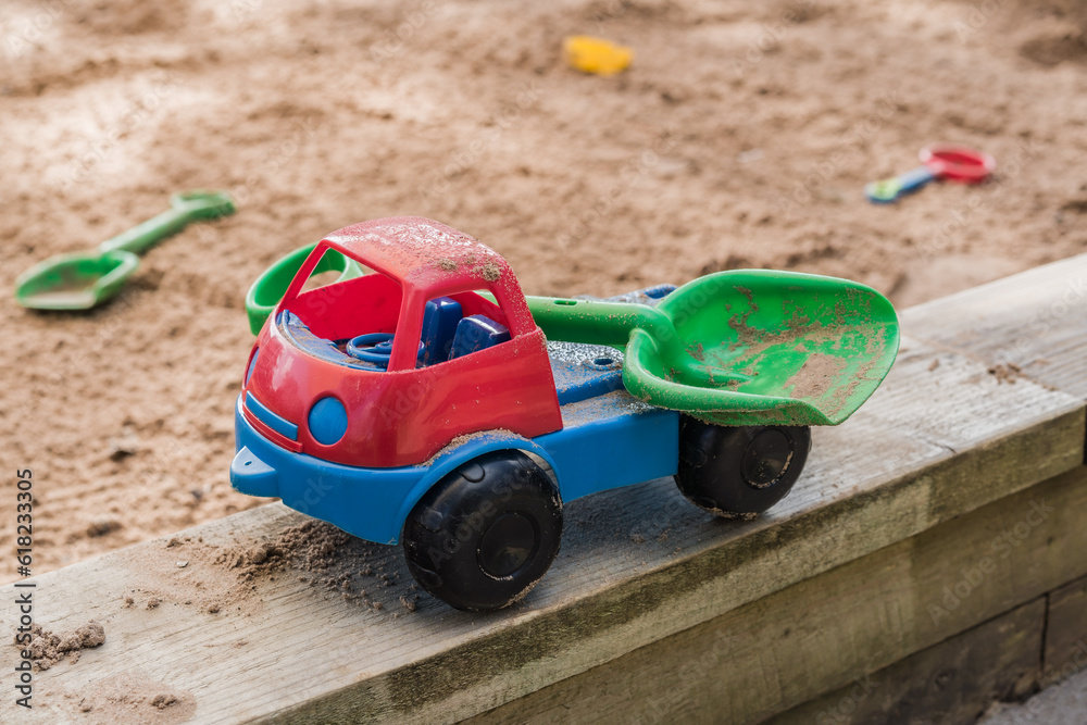 Red and blue toy truck carrying green shovel against sand pit in background, leisure and lifestyle concept illustration. Red and blue toy truck carrying green shovel against sand pit in background, leisure and lifestyle concept illustration.