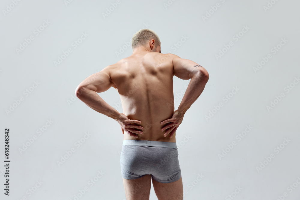 Rear view image of young man with muscular, relief, fit, strong healthy body, back, posing in underwear shirtless against grey studio background. Concept of male beauty, body care, health, sport, ad