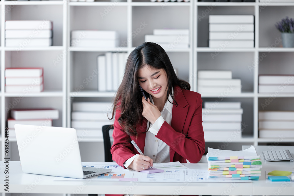 Beautiful asian businesswoman working on laptop and talking the phone. Successful business results in modern office wear red shirt. Beautiful asian businesswoman working on laptop and talking the phone. Successful business results in modern office wear red shirt.