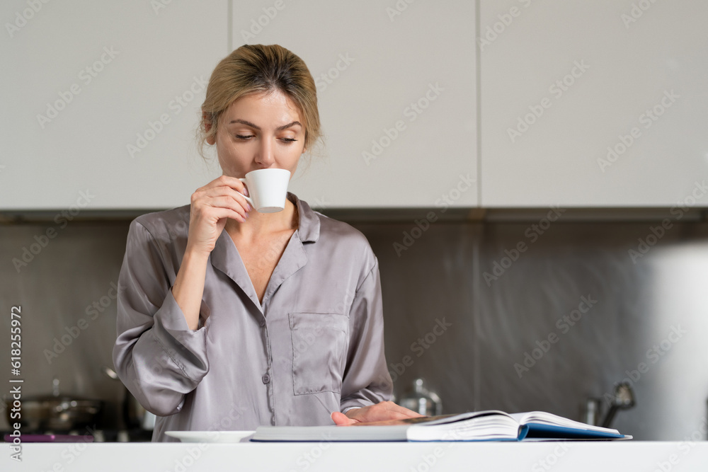 Sexy woman drink coffee and reading book or magazine in the kitchen. Woman wearing pajama relaxing in home kitchen.