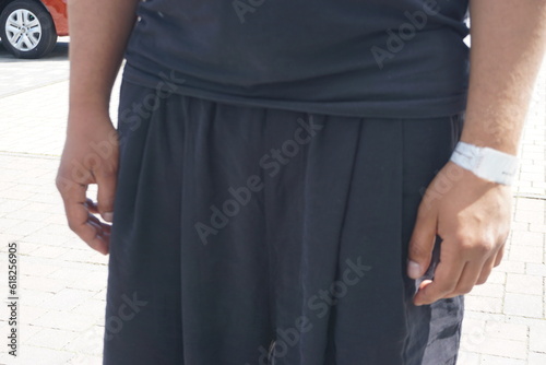 Mann mit schwarzem T-Shirt, schwarzer Pumphose und weißem Verband um Handgelenk steht auf Parkplatz von Krankenhaus bei Sonne am Mittag im Sommer