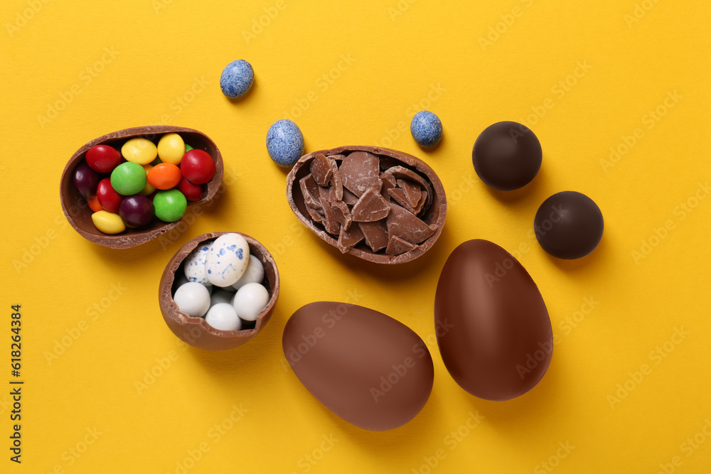 Whole and halves of chocolate eggs with colorful candies on yellow background, flat lay