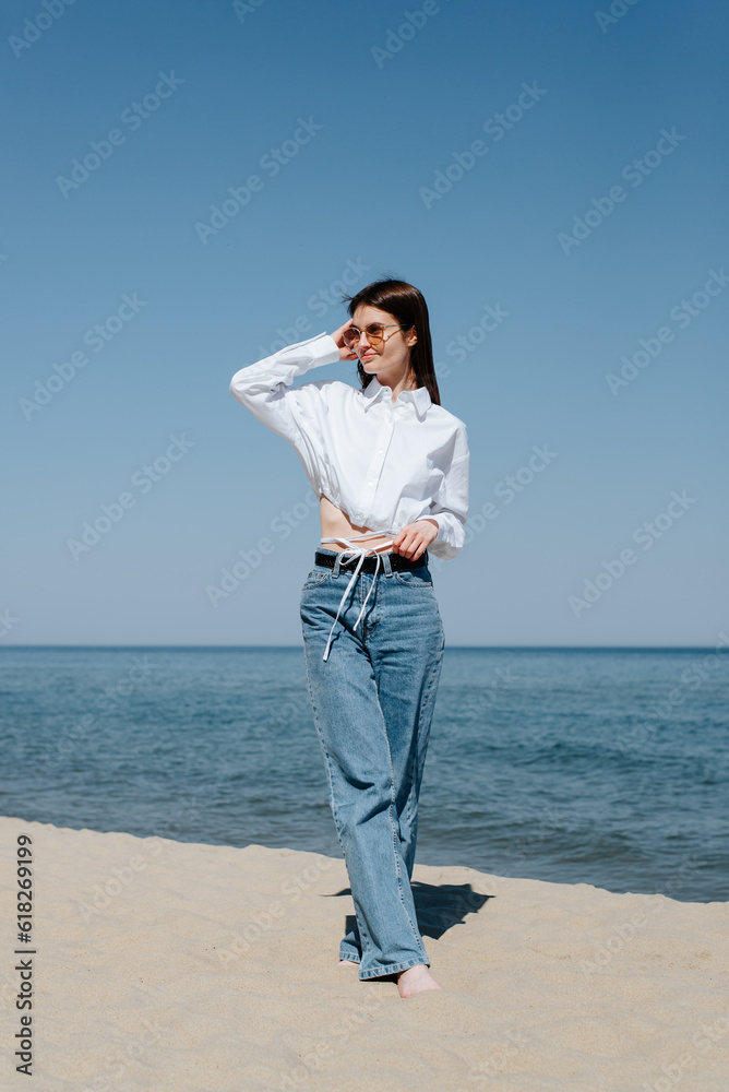 Smiling caucasian young woman in sunglasses on the beach on a sunny day, slim barefoot woman standing on the beach on the sand looking away