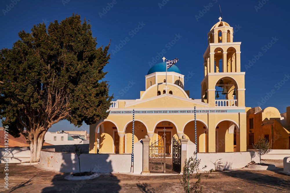 Saint Georgios Oia Holy Orthodox Yellow Church with its Bells Tower and Blue Dome - Oia Village, Santorini Island, Greece